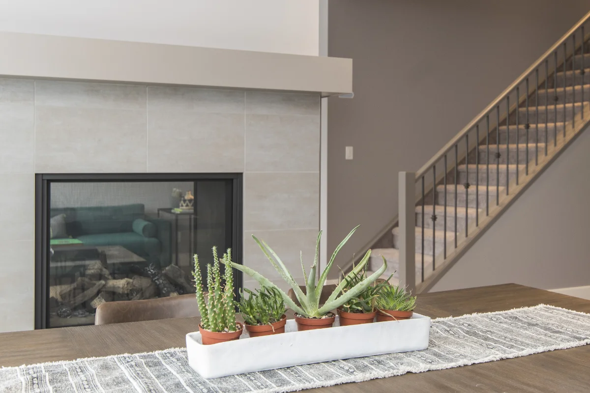 home-img-3 Row of potted succulents in a white planter on a wooden table in a modern living room with a fireplace and staircase.